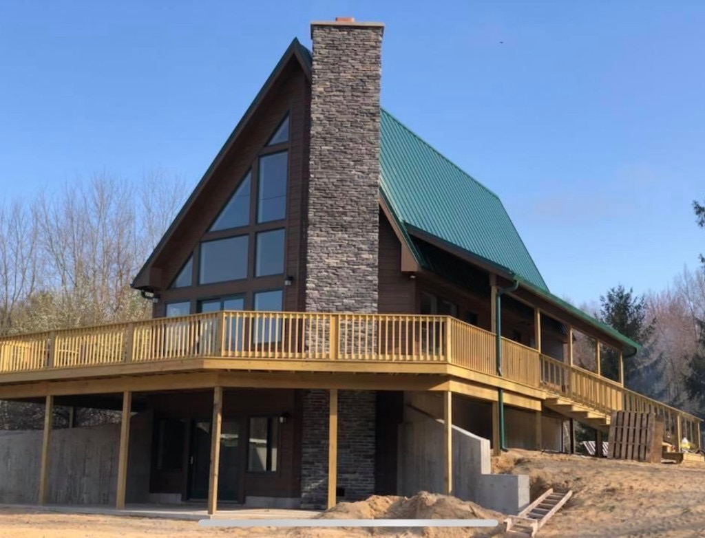 Two-story cabin with green metal roof, stone chimney, and wrap-around deck